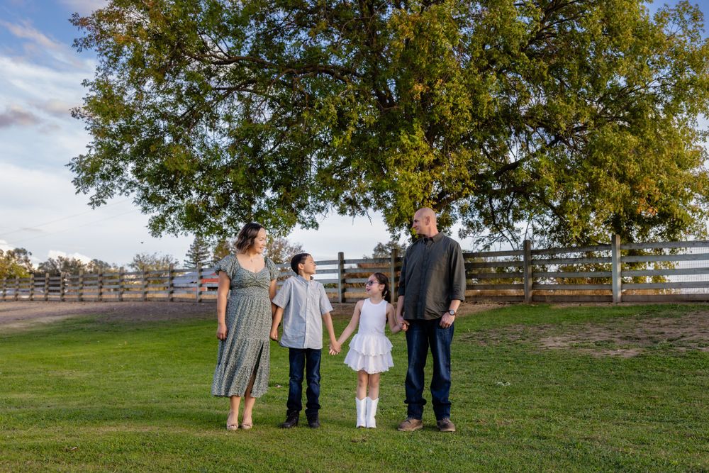 Family of four poses together on grassy field with wooden fence in background.