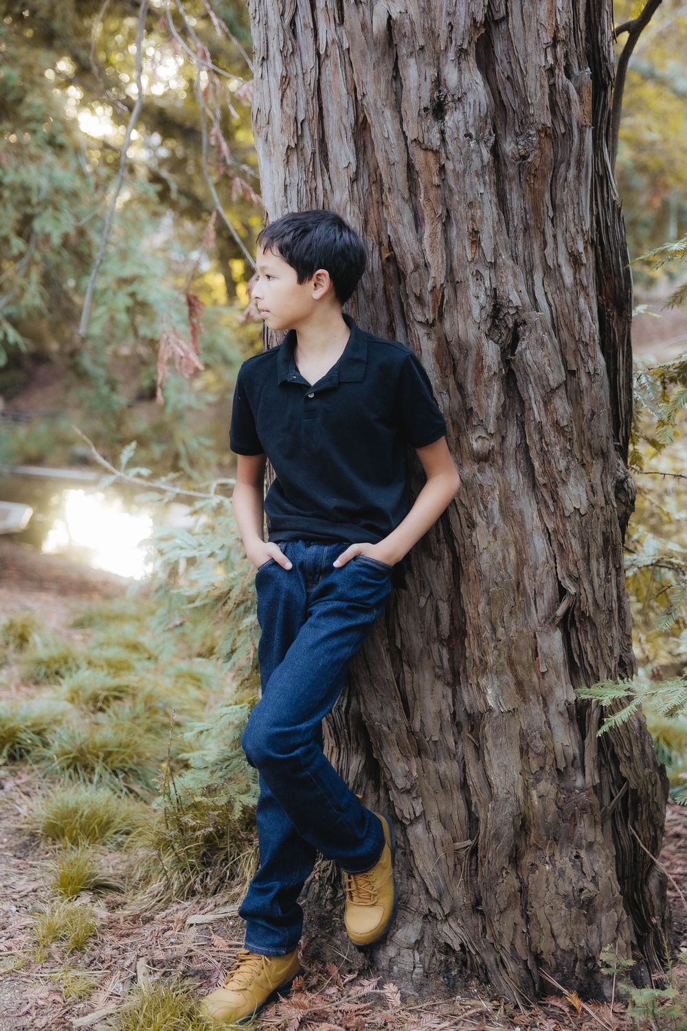 A person in a black polo and jeans leans against a large tree trunk in a wooded outdoor setting.