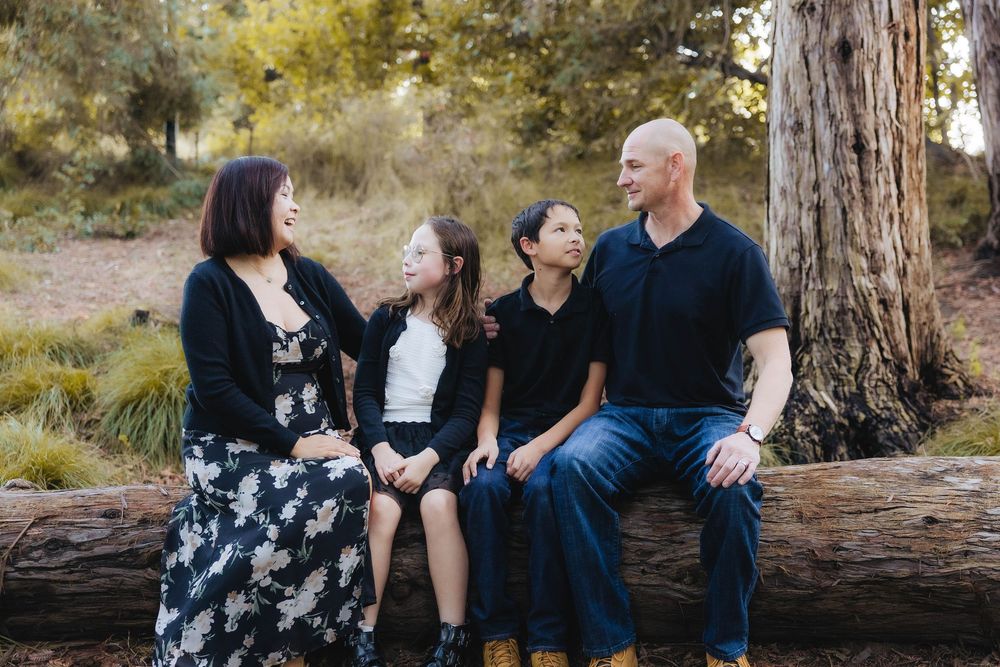 A family sits together on a fallen log in a wooded outdoor setting during a casual photo session.
