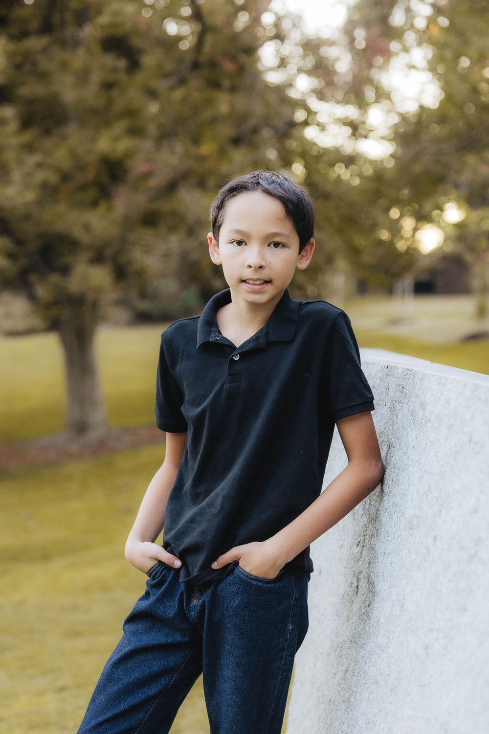A child in a navy blue outfit poses casually against a white wall outdoors during golden hour.