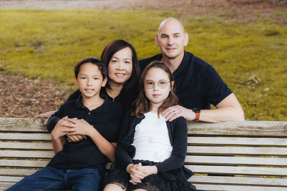 A family sitting together on a wooden bench outdoors in casual clothing against a natural background.