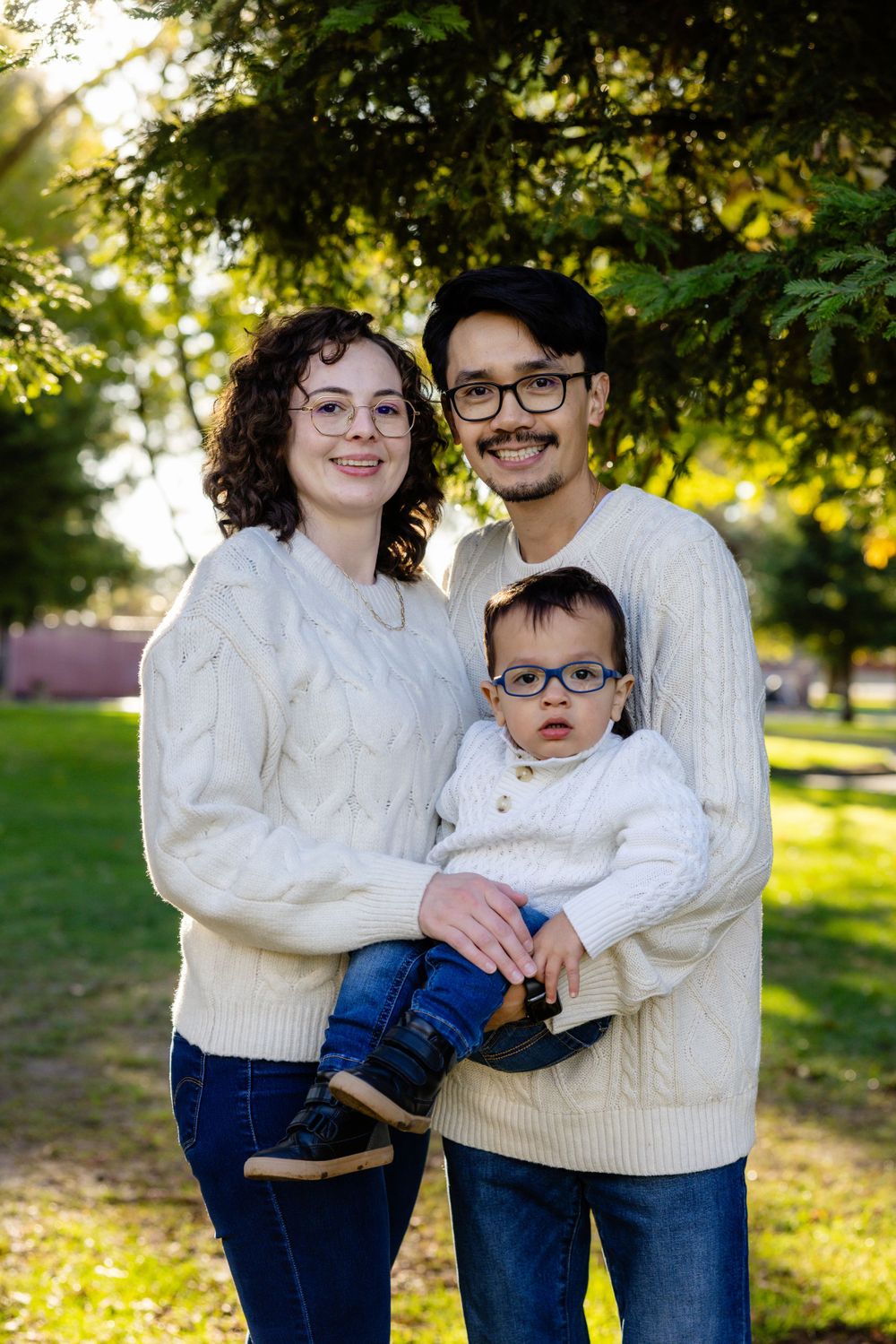 Family of three in matching cream sweaters pose for portrait in sunny park setting.