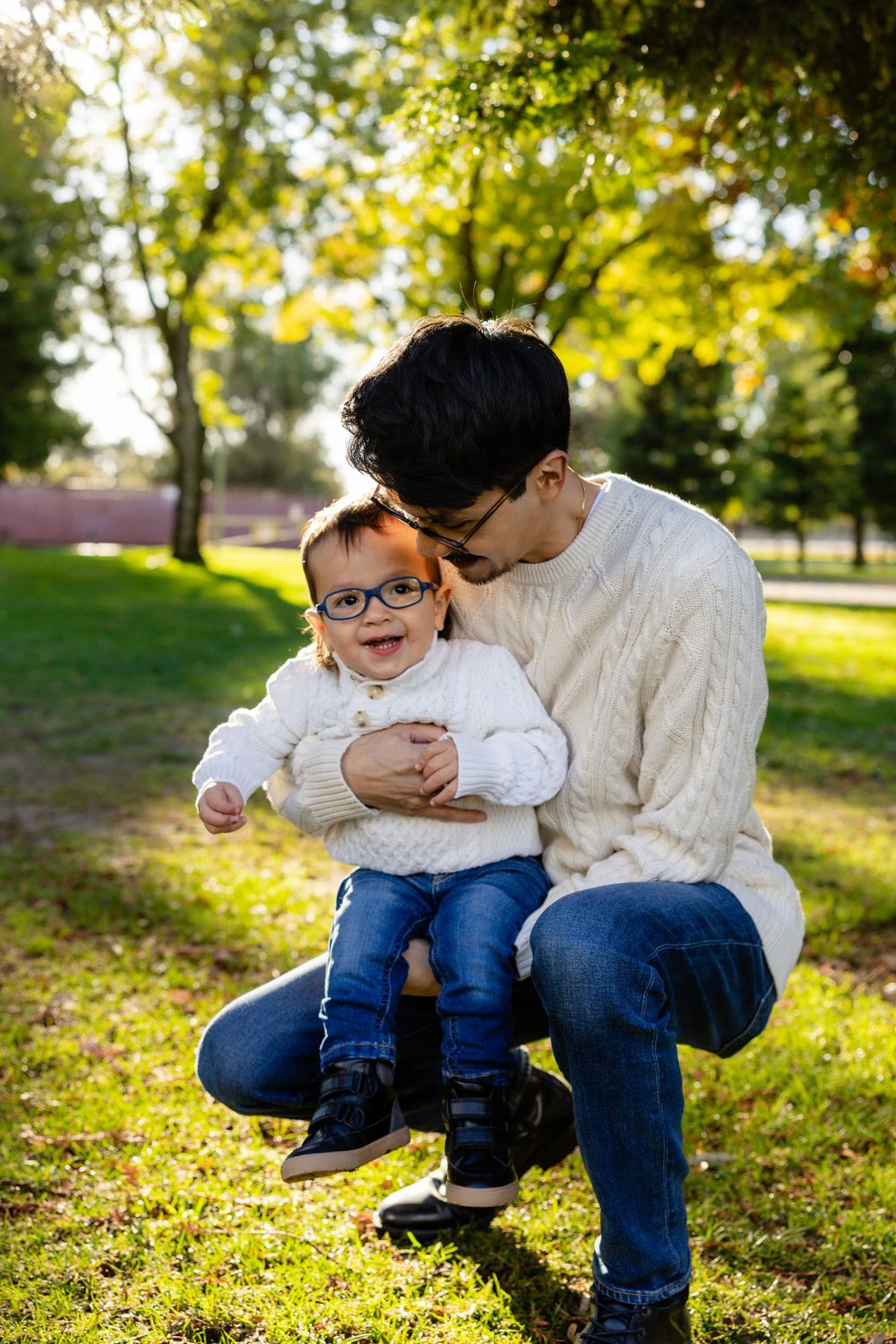 Parent and child in matching white sweaters share tender moment in sunlit park.