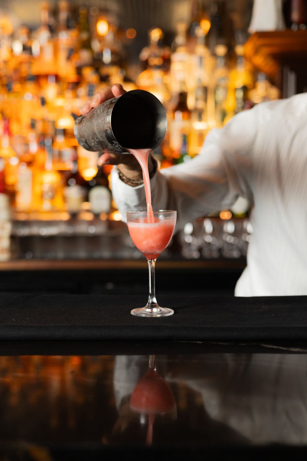 Pink cocktail being strained from a shaker into a martini glass at an elegant bar.
