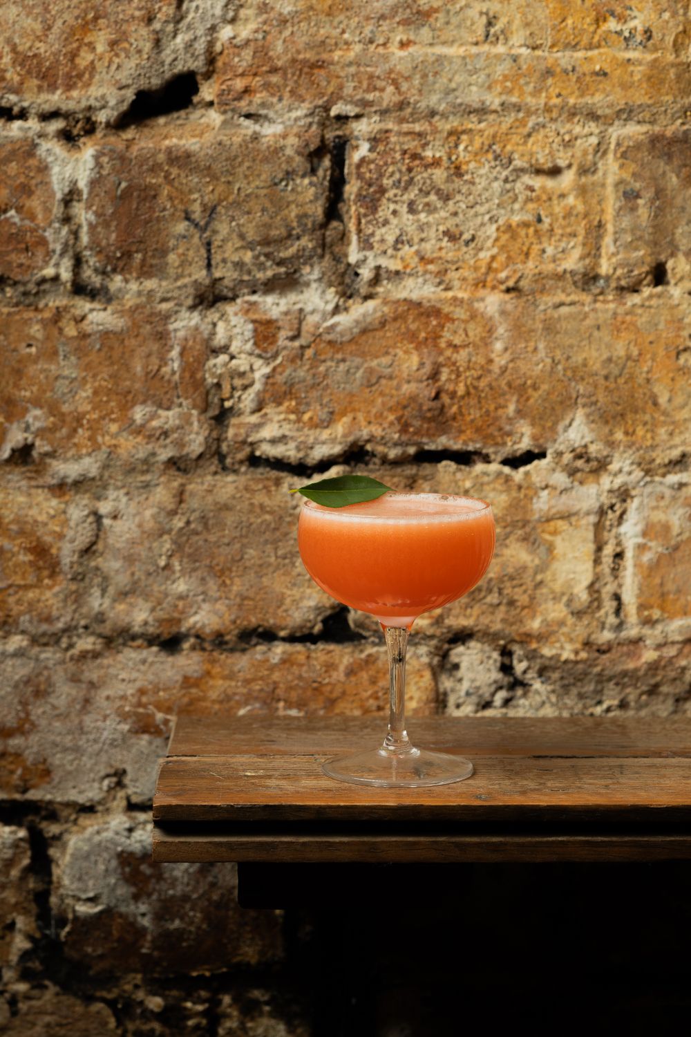 Orange-colored cocktail in coupe glass against rustic exposed brick wall.