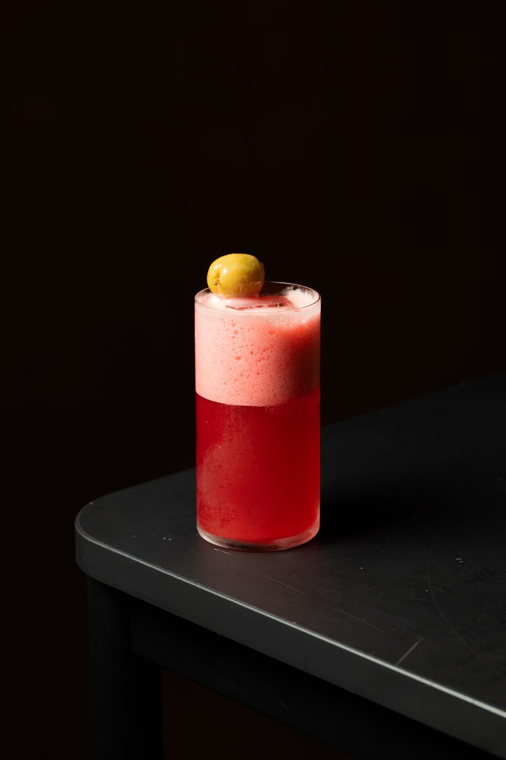 Red layered cocktail with pink foam and olive garnish on black surface.