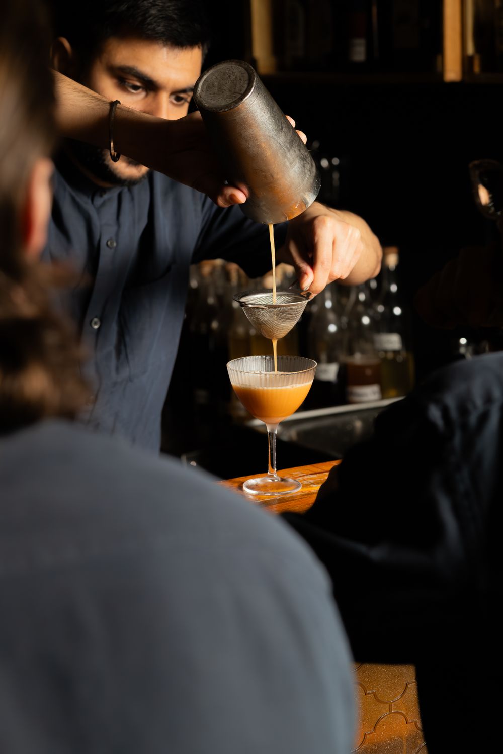 Bartender pouring a cocktail from a shaker into elegant martini glasses at a dimly lit bar.