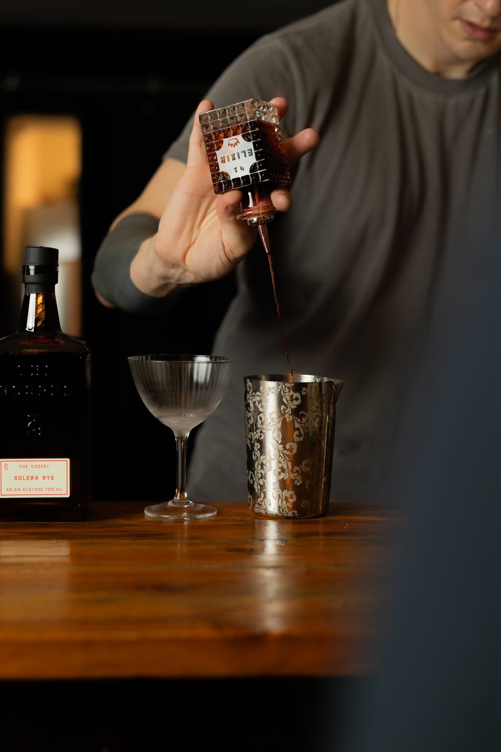 Pouring bitters into a cocktail shaker with bar tools and glassware on wooden surface.