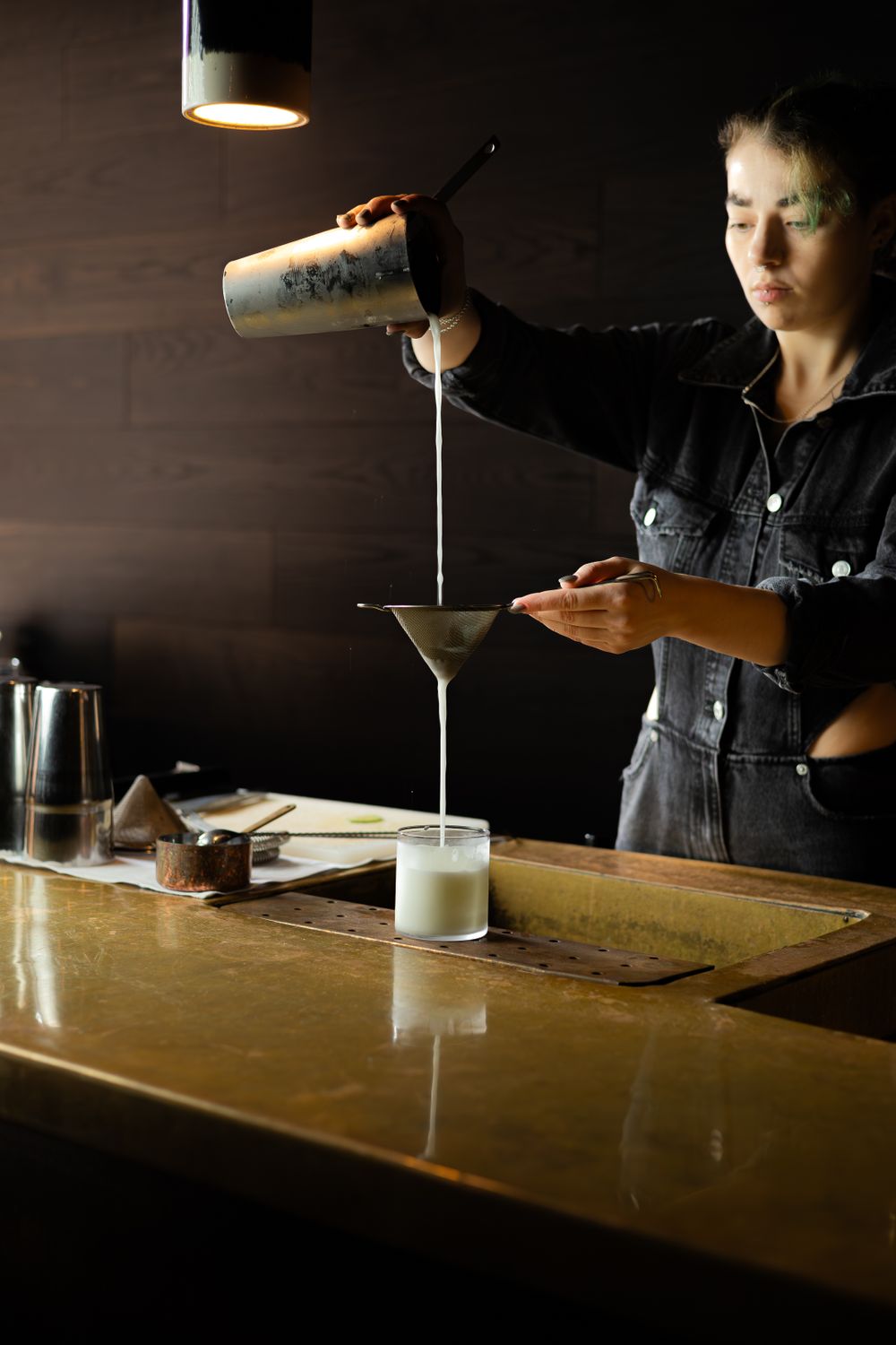 Bartender pouring white liquid from shaker into glass on wooden bar counter.