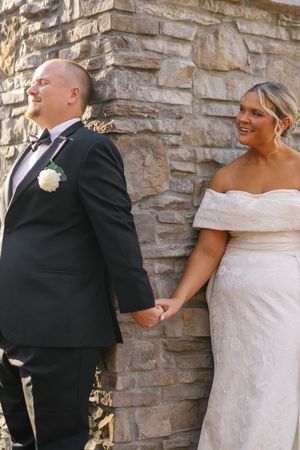 A couple shares a joyful moment on their wedding day while standing against a stone wall.