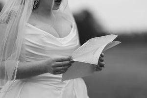 A bride in a white wedding dress reads a letter or note while wearing a veil in this black and white photograph.
