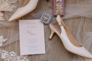 Wedding accessories including white bridal shoes, invitation cards, and decorative elements arranged on a wooden surface.