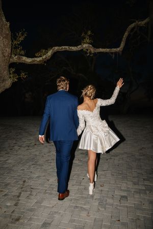 A couple in wedding attire seen from behind walking on brick pavement at night under a twisting tree branch.