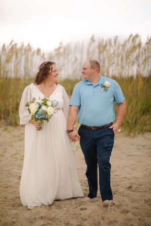 A couple walks hand in hand on a sandy beach with sea grass in the background during a casual beach wedding.