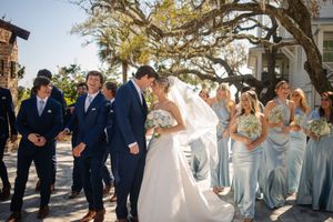 Wedding party in navy suits and light blue dresses celebrate with couple under sunlit oak tree branches outdoors.