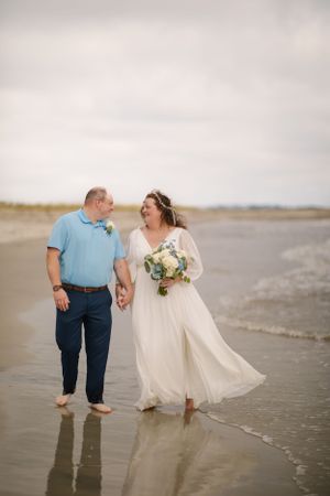 A couple shares a joyful moment walking barefoot along a beach shoreline at sunset with wedding attire and flowers.