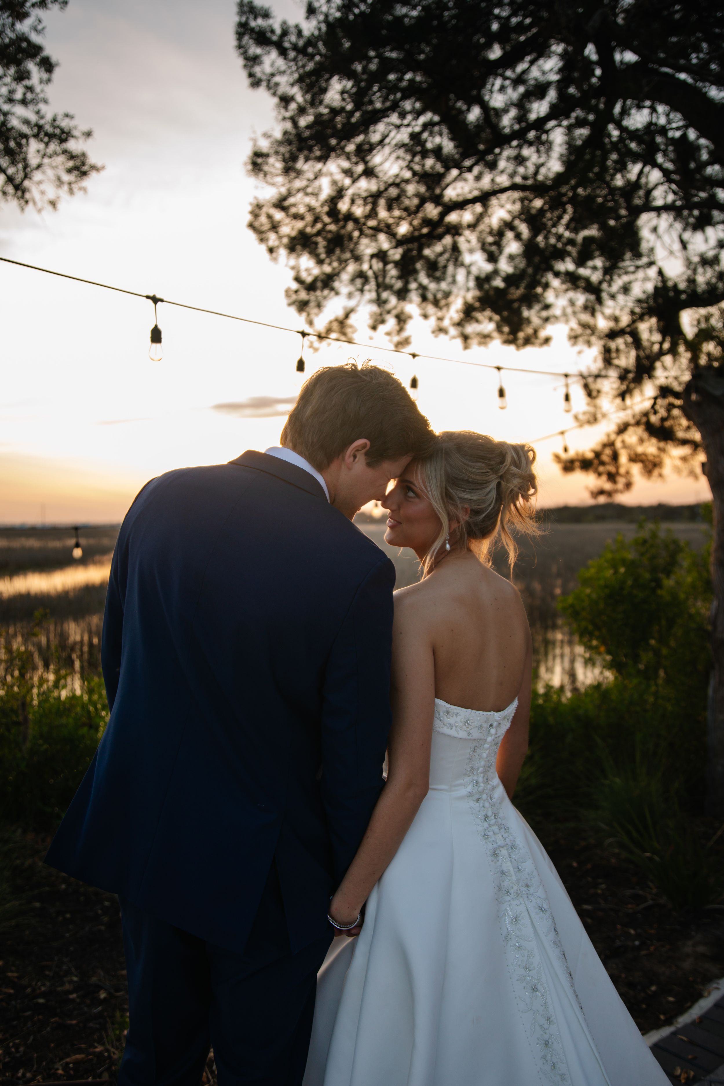 A couple shares an intimate moment at sunset with string lights overhead and marsh views in the background.