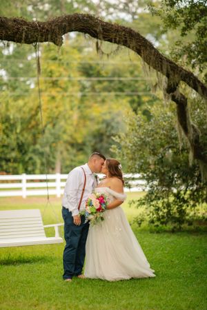 A romantic wedding couple embraces under a moss-draped oak tree branch on a grassy lawn with white fencing in the background.