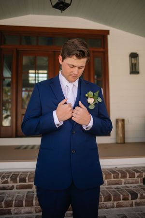 A groom in a navy blue suit with a white boutonniere adjusts his jacket while standing on brick steps.
