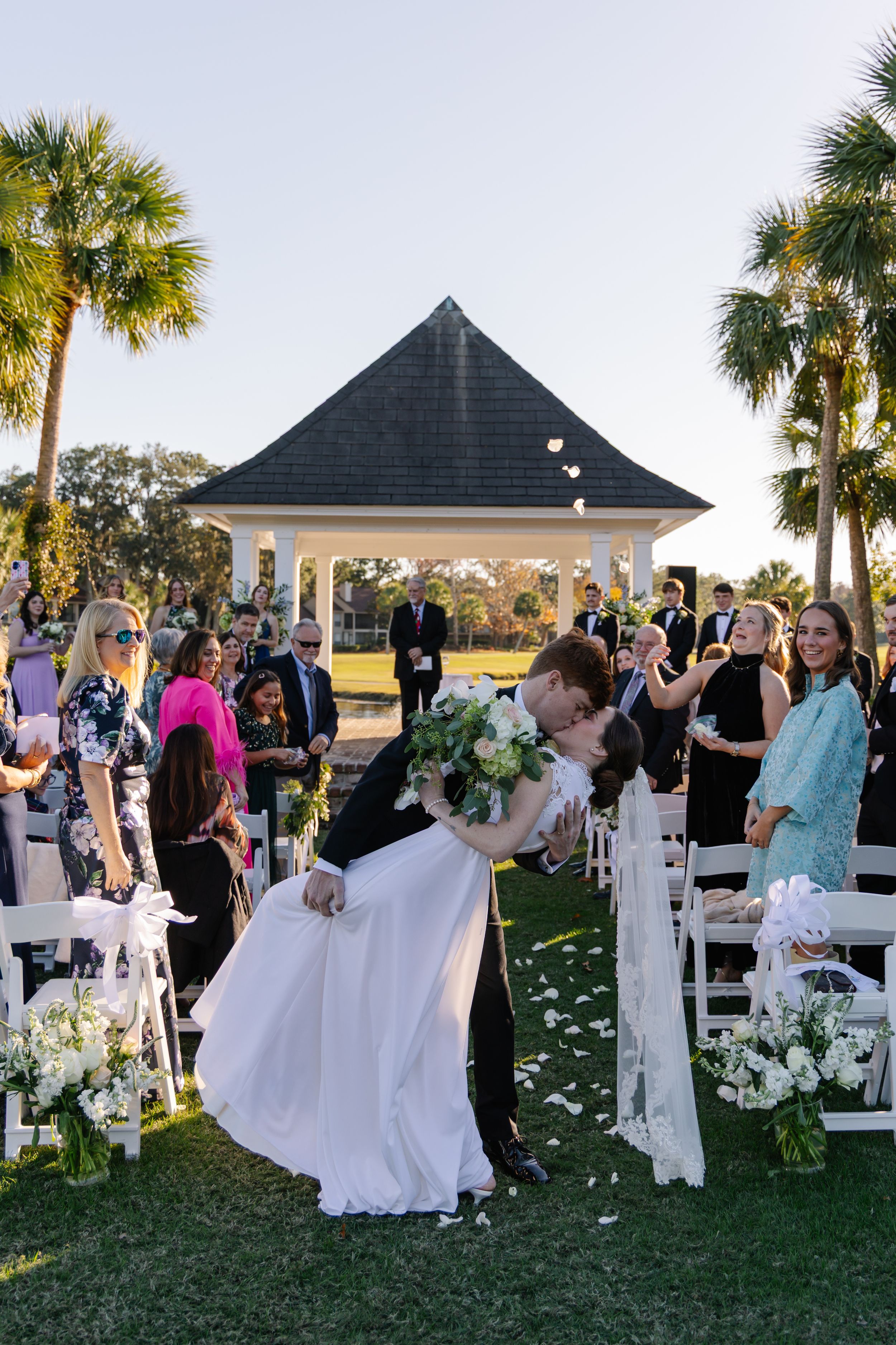 Wedding guests dance and celebrate during an outdoor reception at sunset with palm trees and a white gazebo in the background.