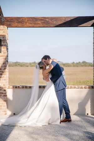 A romantic outdoor wedding moment with a couple in formal attire embracing under a rustic wooden beam structure.