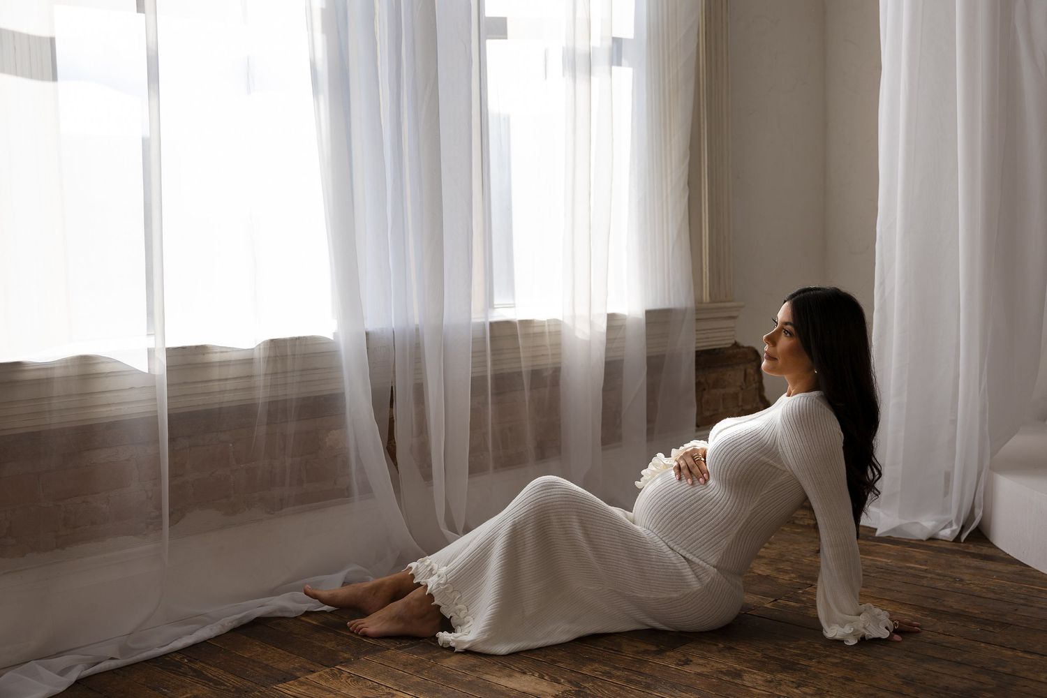 White sheer curtains billow in the breeze by a window, creating a soft and ethereal atmosphere in the room with pregnant women