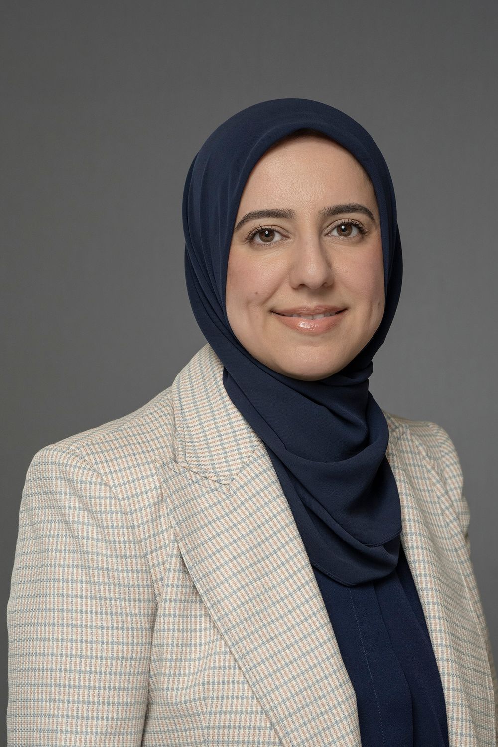 Professional woman wearing navy hijab and checkered blazer posing for corporate headshot on gray background.