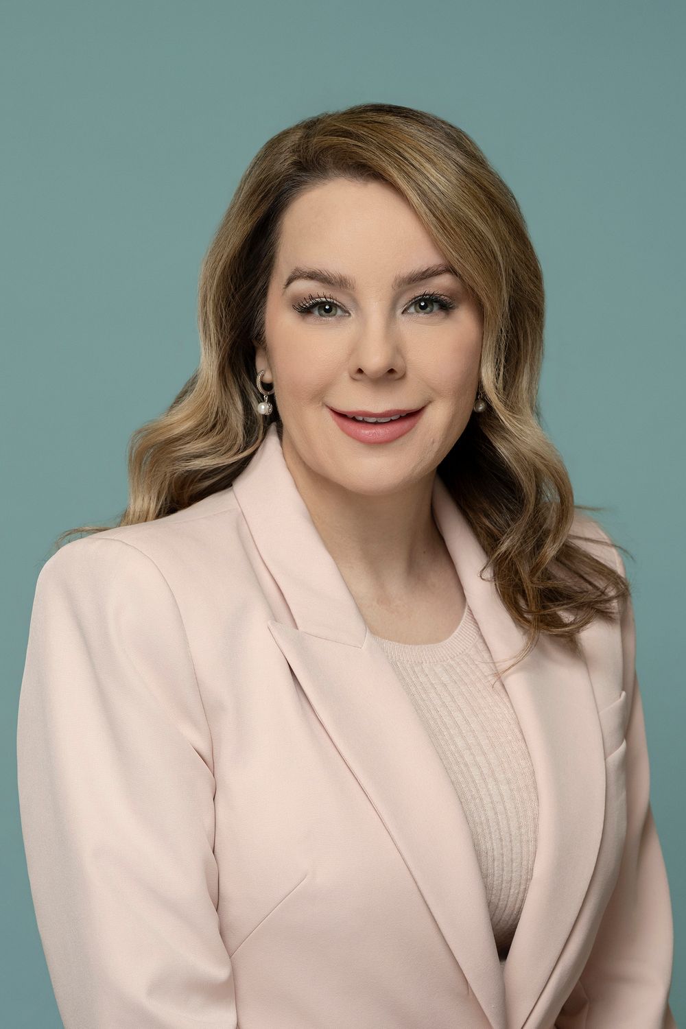 Elegant woman with wavy blonde hair in pink blazer posing for professional portrait on teal background.