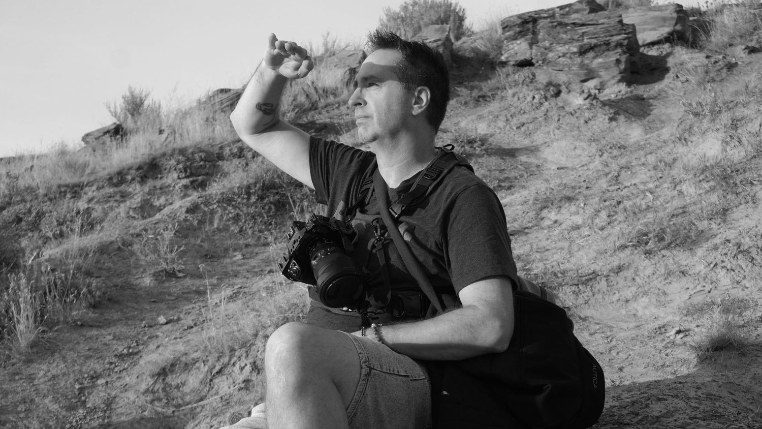A person in black clothing and a hat walks along a rocky desert trail in black and white.