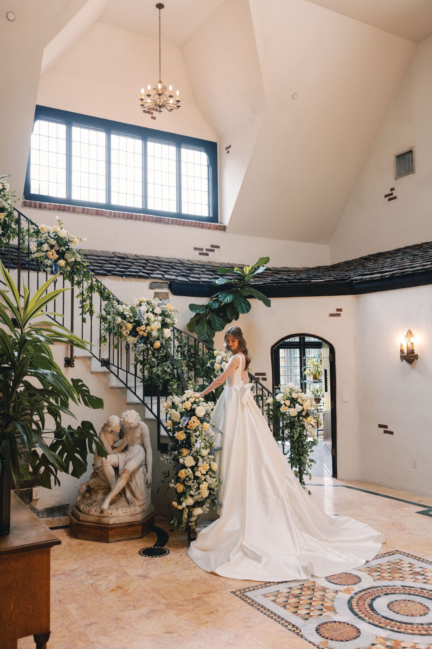 Bride in flowing white gown standing on grand staircase adorned with floral arrangements.