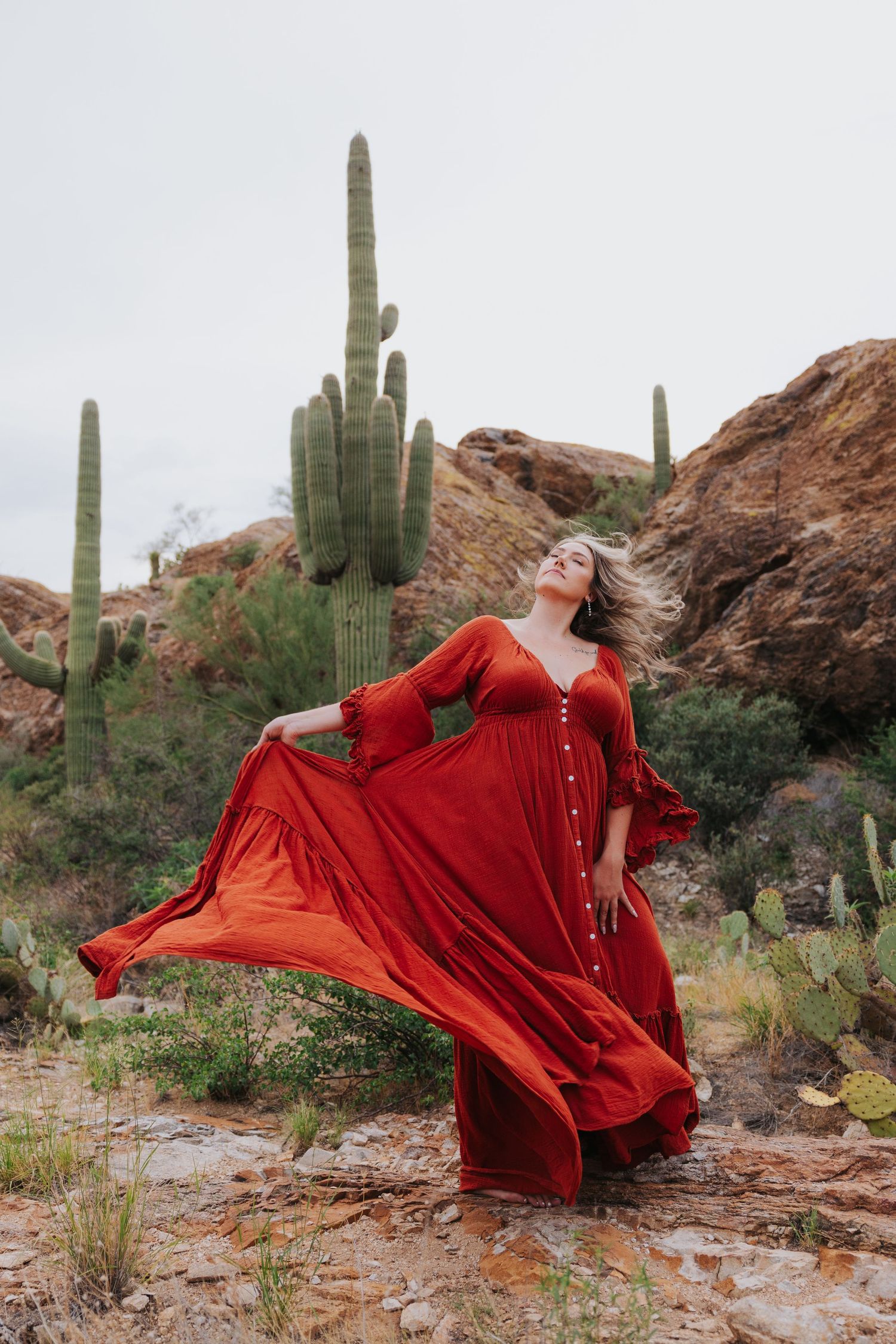 A flowing red dress creates dramatic movement against a desert landscape with tall saguaro cacti and rocky terrain.