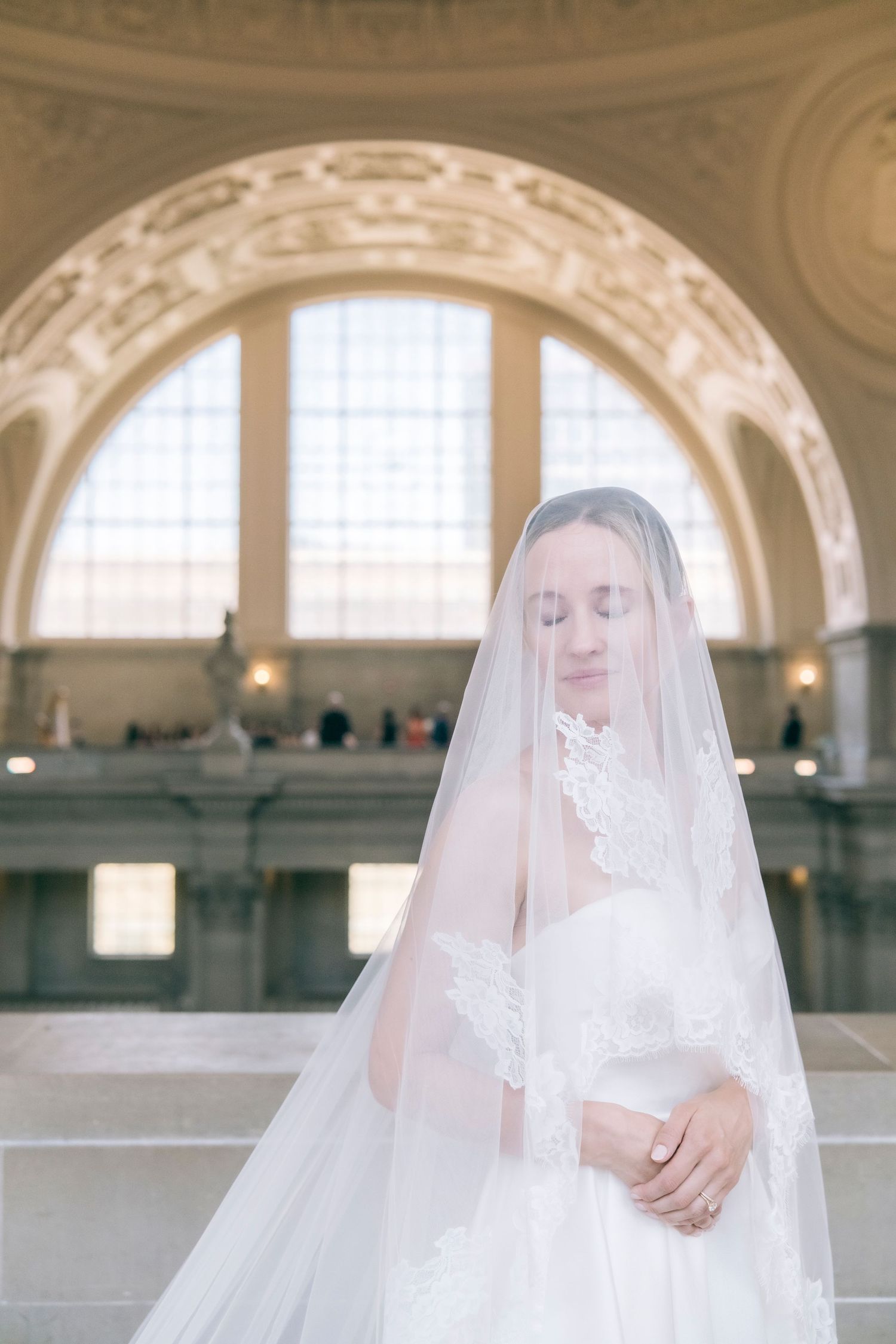 Serene bride in lace-trimmed veil with eyes closed under grand arched ceiling of city hall.
