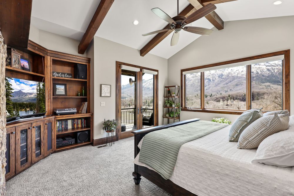 Master bedroom with vaulted beam ceiling, ceiling fan, built-in shelving, and panoramic mountain view windows with balcony access.
