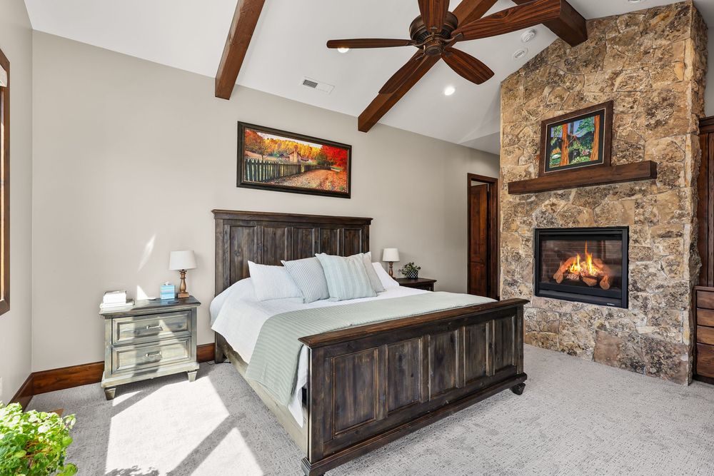 Cozy bedroom featuring dark wood bed frame, vaulted ceiling with beams, and a stone fireplace with wooden mantel. Captured by Slate Media