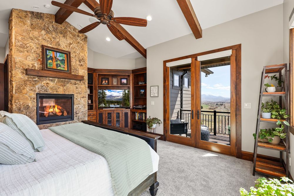 Spacious master bedroom with vaulted ceiling, stone fireplace, built-in shelving, and French doors leading to a scenic balcony.