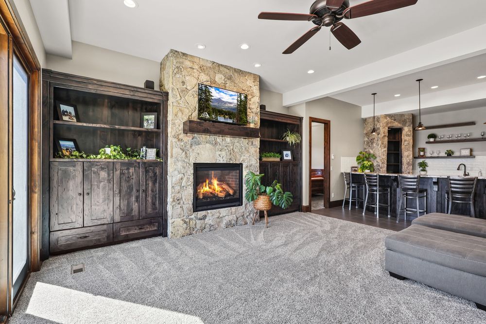 Inviting living room with stone fireplace, built-in dark wood cabinets, ceiling fan, and open concept dining area. Captured by Slate Media