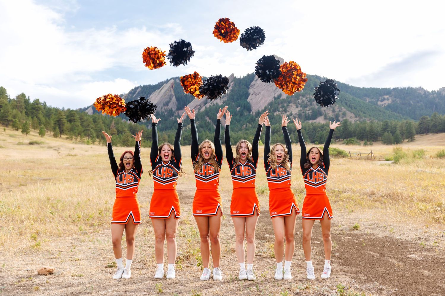 Six cheerleaders in orange uniforms toss colorful pom-poms in the air outdoors with mountains in the background.