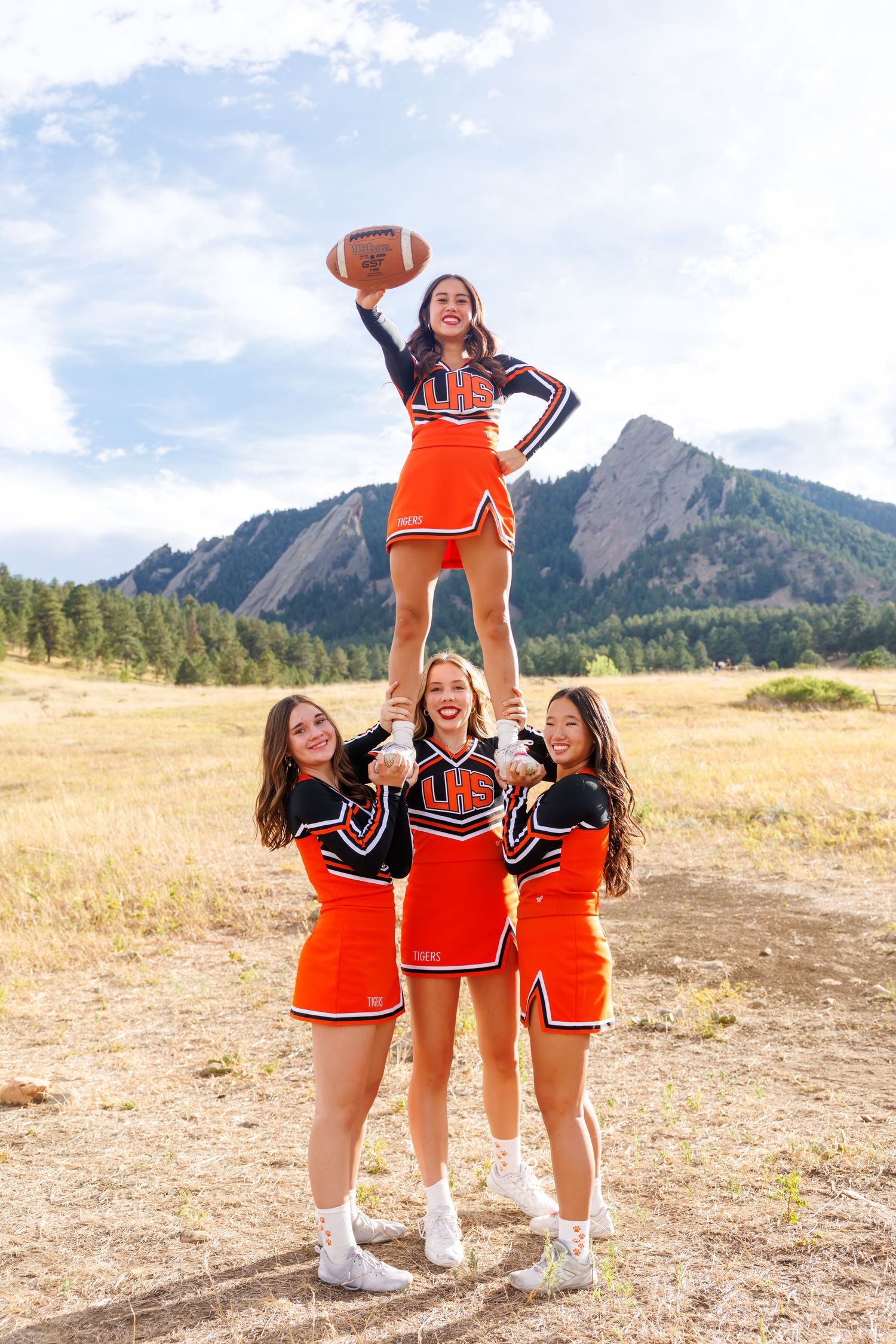 Four cheerleaders in orange uniforms perform a stunt outdoors near the Flatirons in Boulder, Colorado, holding a football.