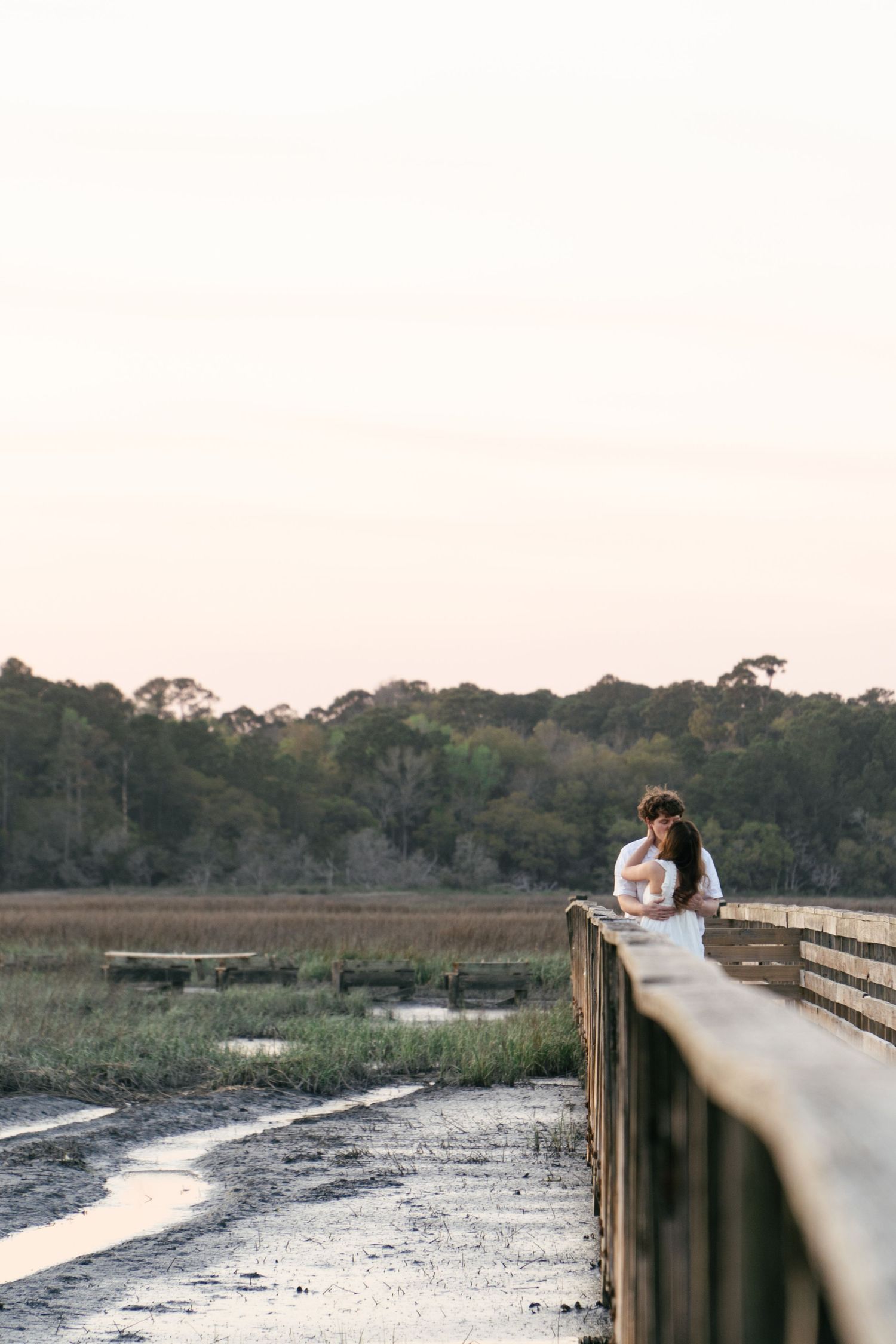 A scenic panoramic view of a wooden boardwalk winding through wetlands at sunset with reflective water.