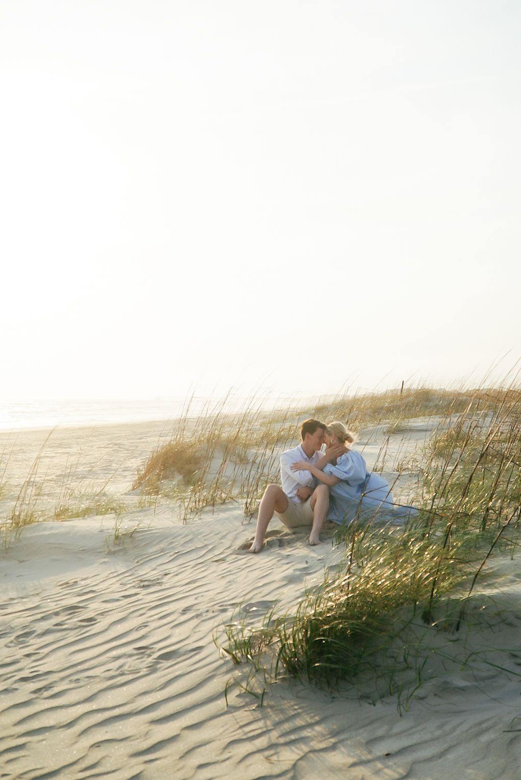 A couple sits together on a sandy beach dune with beach grass at sunset, creating a romantic coastal scene.