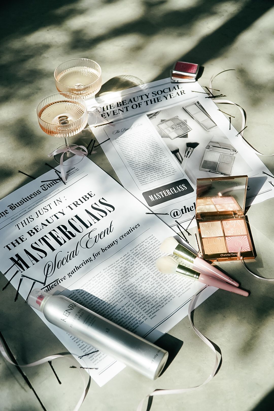 A flatlay of beauty products with champagne glasses, makeup tubes, and newspaper articles on a sunlit surface.