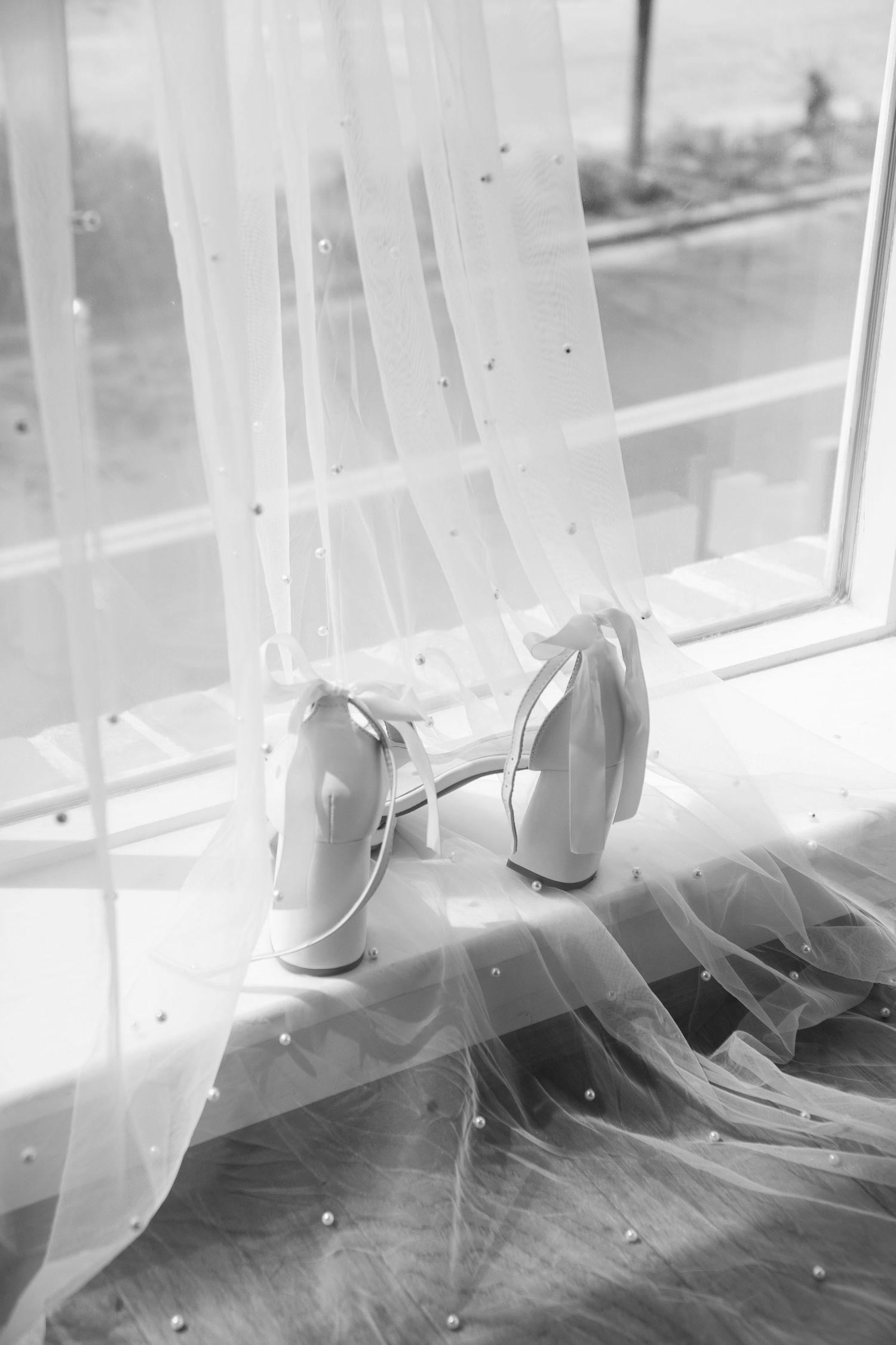 A pair of elegant white bridal shoes next to a sheer curtain by a window in black and white photography.