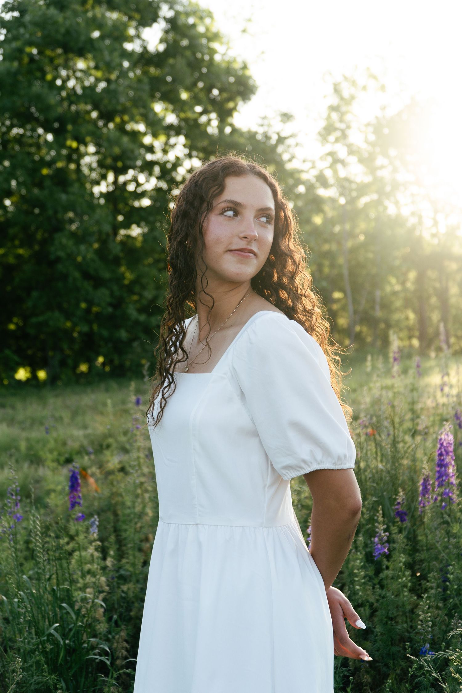 A white dress flows gracefully in a field of purple flowers during a sunset photoshoot.
