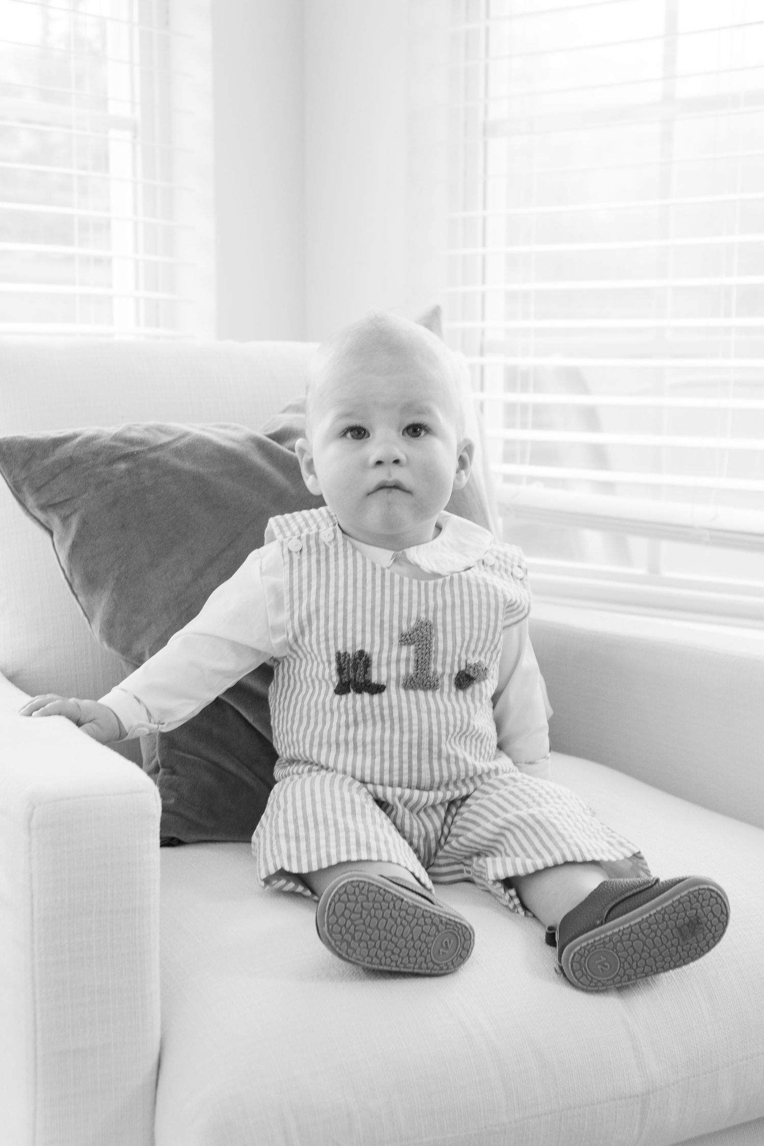 A small child sits on a white couch wearing striped overalls and gray shoes in a black and white photograph.