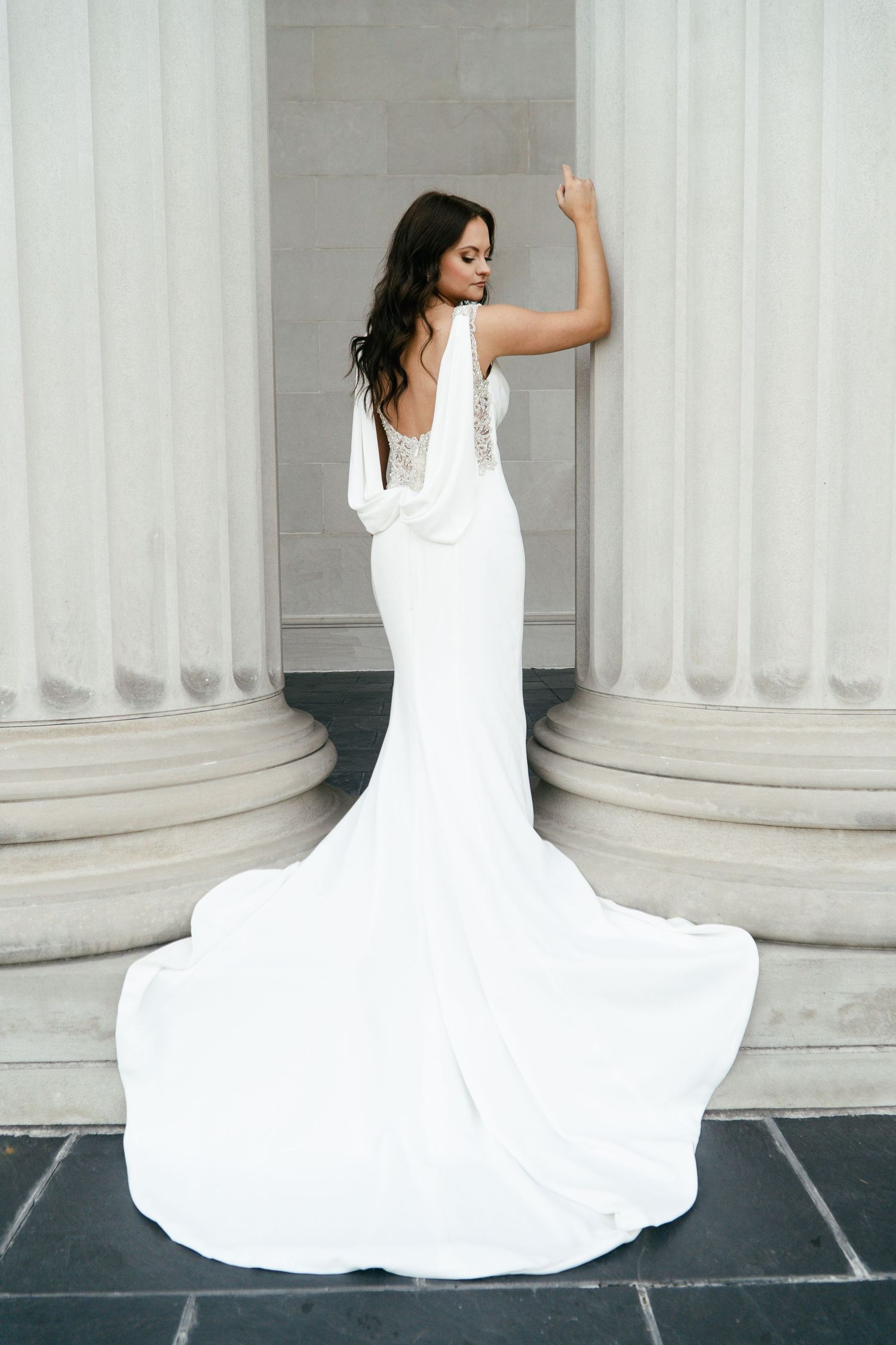 A bride in an elegant white mermaid wedding dress poses between classical columns with a flowing train.
