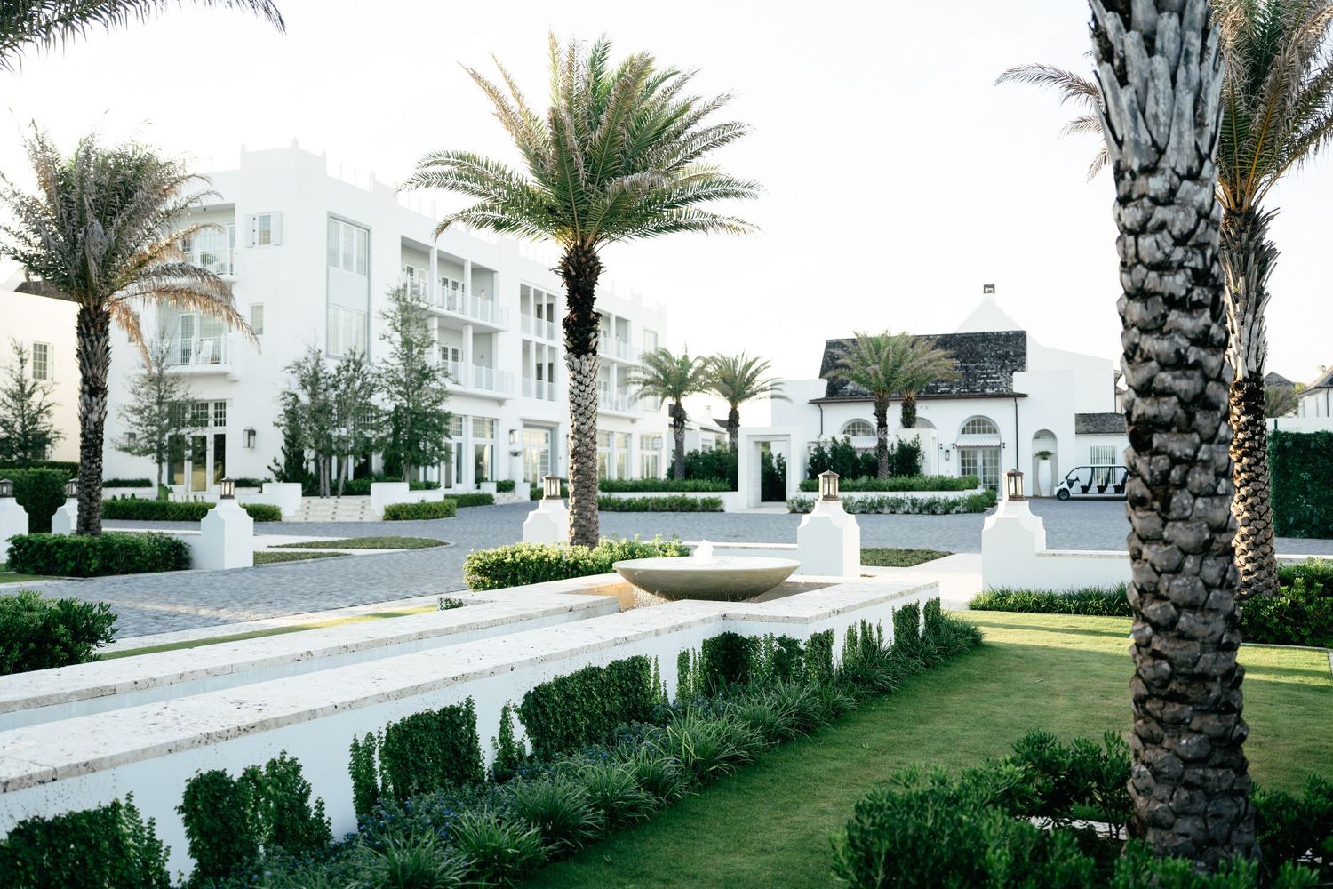 Palm trees line a curved walkway with white walls and buildings in a modern residential complex.