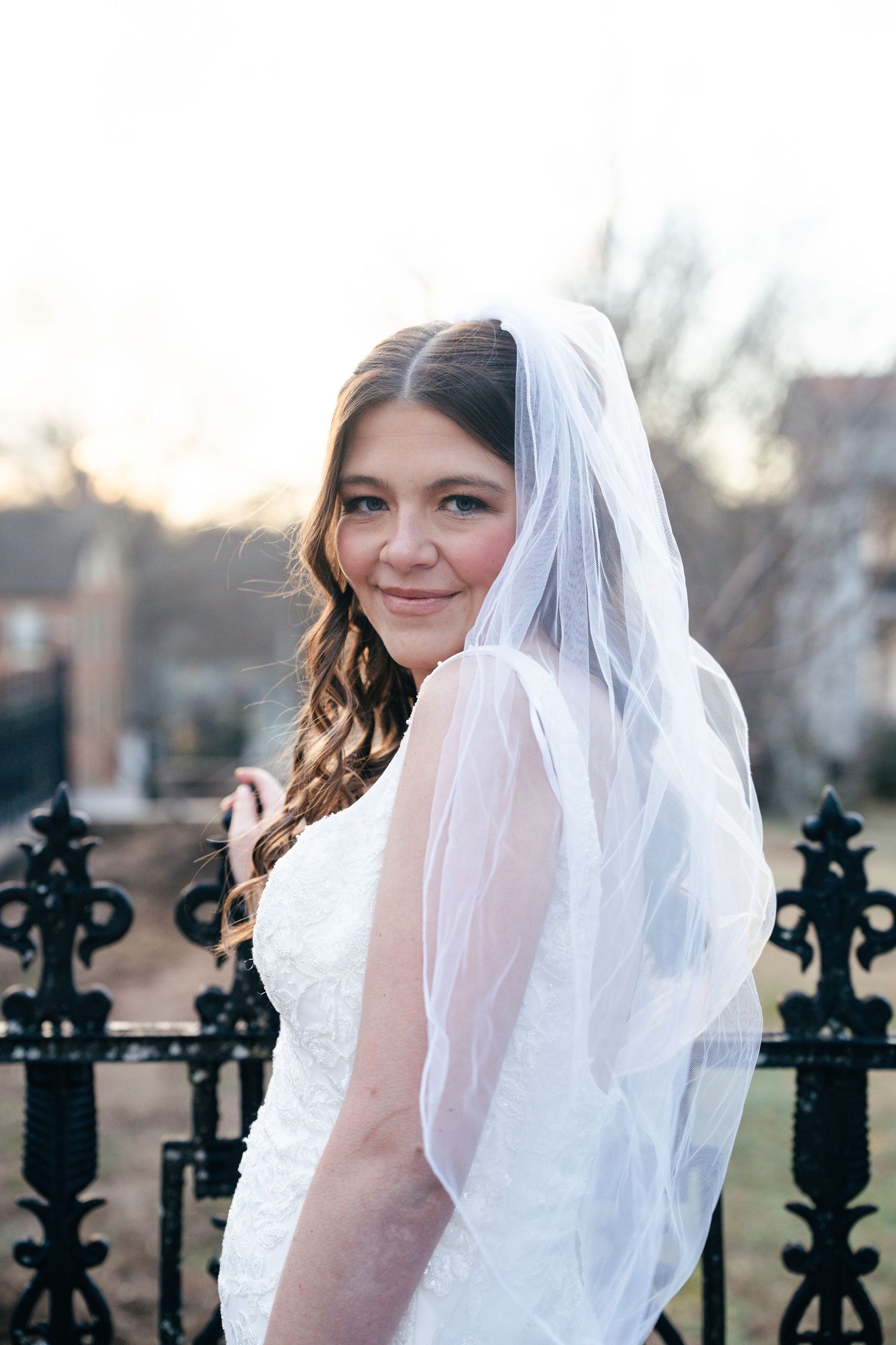 Smiling bride in white lace gown and veil poses by ornate black iron fence outdoors on her wedding day.