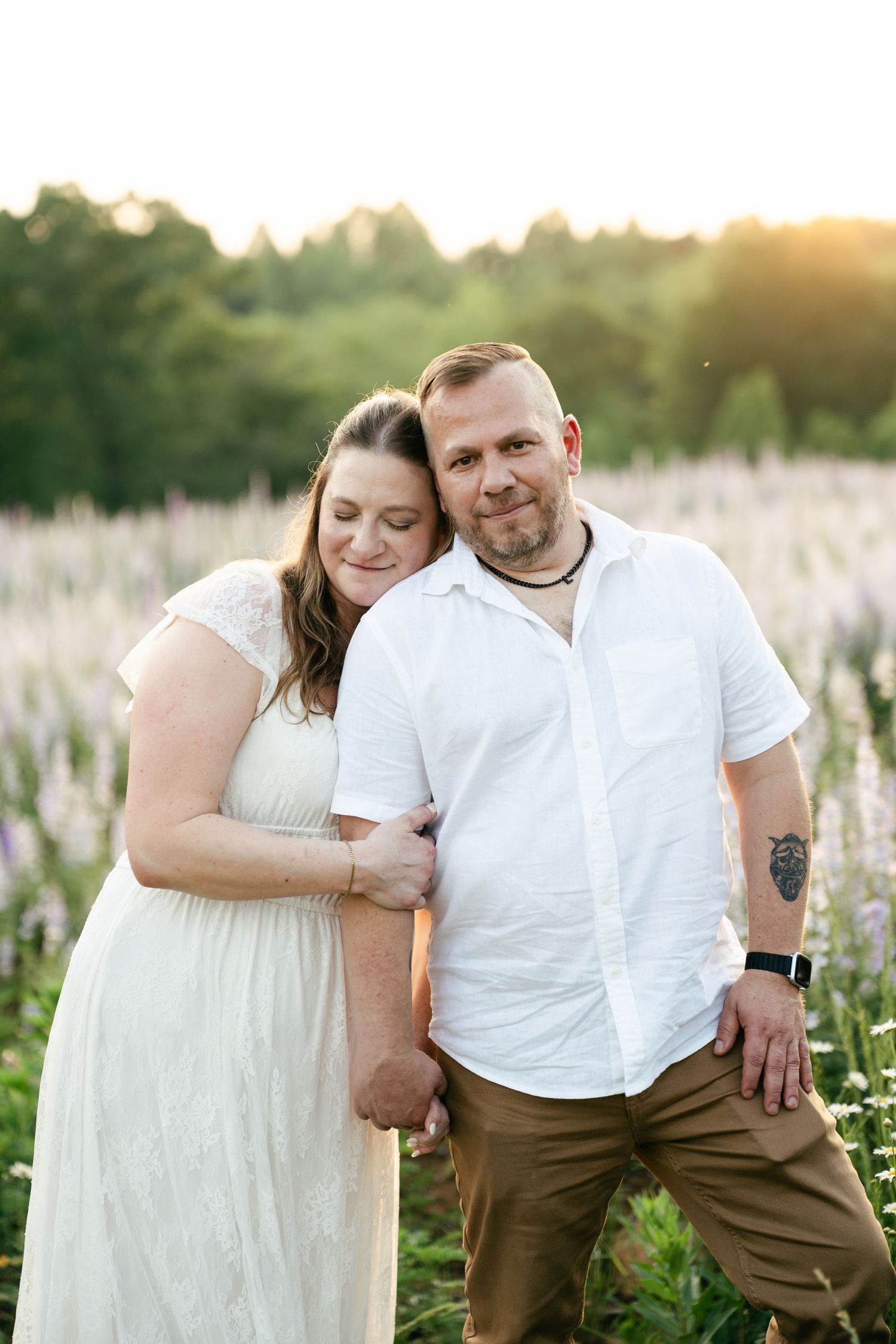 A couple in white outfits embraces while standing in a wildflower meadow at sunset.