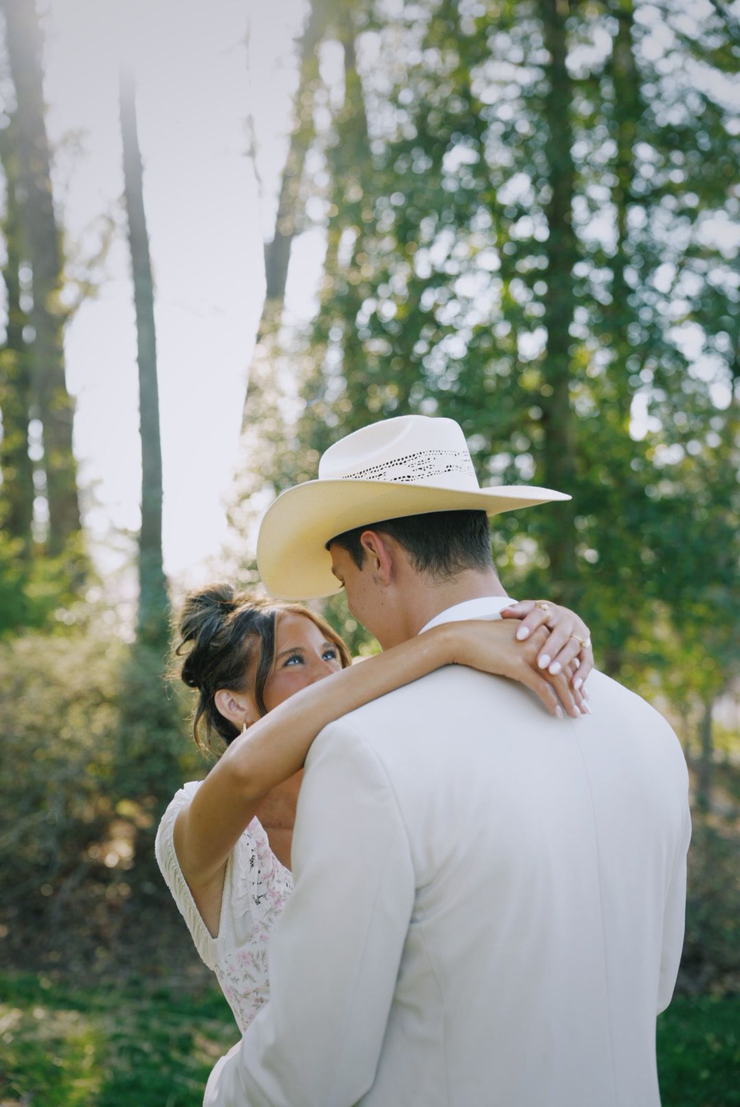 A bride and groom embrace outdoors, groom wearing a white cowboy hat and suit, surrounded by lush green trees.