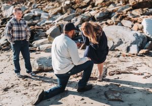 A romantic beach proposal moment with people positioned on rocky coastal terrain during sunset.  Mitchell Field in Harpswell, ME.
